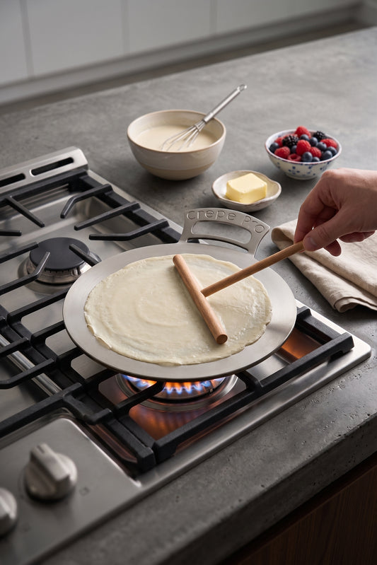 A hand uses a wooden spatula to flip a thin crepe on a round carbon steel 12" Crepe Pro pan on stovetop over a gas flame in a kitchen. Bowls of batter, butter, and mixed berries sit nearby on the countertop.