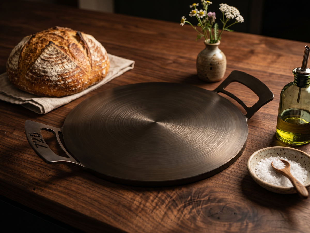 A round dark nitrogen infused carbon steel 14" 726 Pan with side handles sits on a rustic wooden table, styled with a loaf of bread, a small vase of flowers, a bottle of oil, and a bowl of salt.