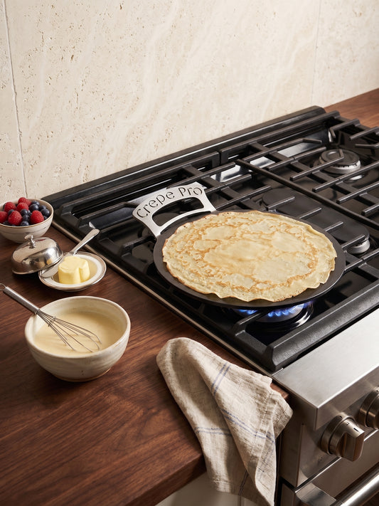 A 12" crepe pro carbon steel pan with a 'Crepe Pro' handle sits on a lit gas stove burner, cooking a thin crepe. The scene is set on a dark wood countertop with bowls of batter, butter, fresh berries, and a checkered kitchen towel, against a textured beige tile wall.
