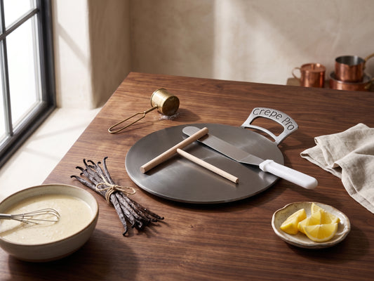 A round dark gray carbons steel 12" crepe pro pan with a 12" crepe spatula with a white handle and a 7" crepe pro crepe spreader, styled on a wooden table with vanilla beans, a bowl of batter, lemon wedges, and kitchen props.