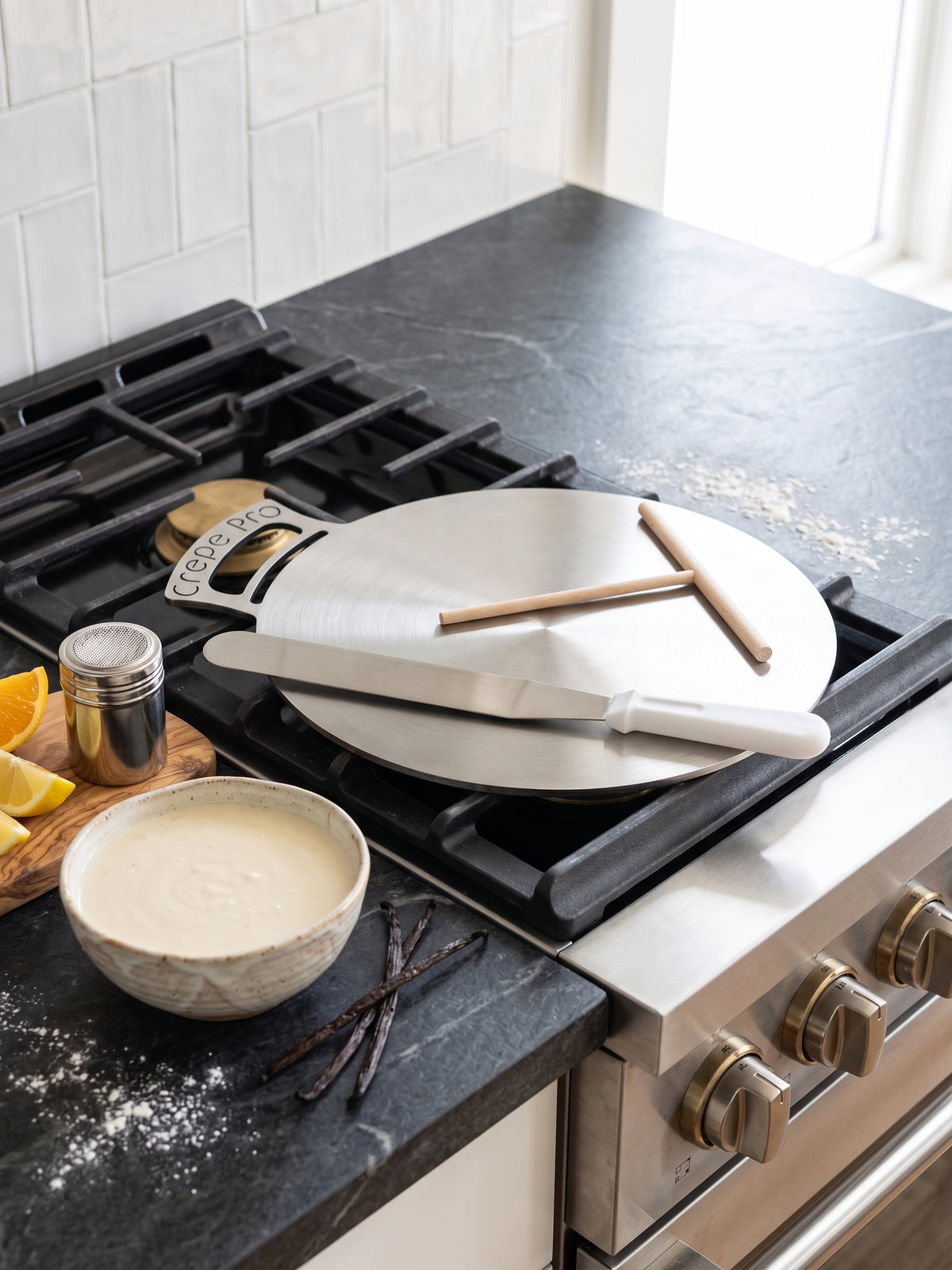 A round carbon steel 16" Crepe pro pan with a 7" crepe spreader and 12" spatula on a black gas stovetop in a kitchen. A metal spatula and two wooden sticks rest on the pan, with a small bowl of batter, vanilla beans, and lemon slices nearby on a dark countertop.
