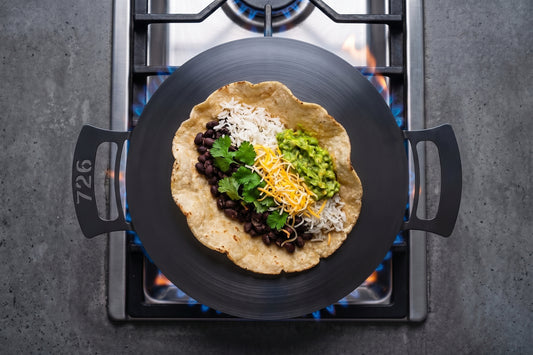 A black nitrogen infused carbon steel 14" 726 pan with side handles sits on a gas stove burner with blue flames, holding a tortilla wrap topped with black beans, shredded white cheese, guacamole, and cilantro.