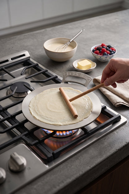 A hand uses a wooden spatula to flip a thin crepe on a round carbon steel 12" Crepe Pro pan on stovetop over a gas flame in a kitchen. Bowls of batter, butter, and mixed berries sit nearby on the countertop.