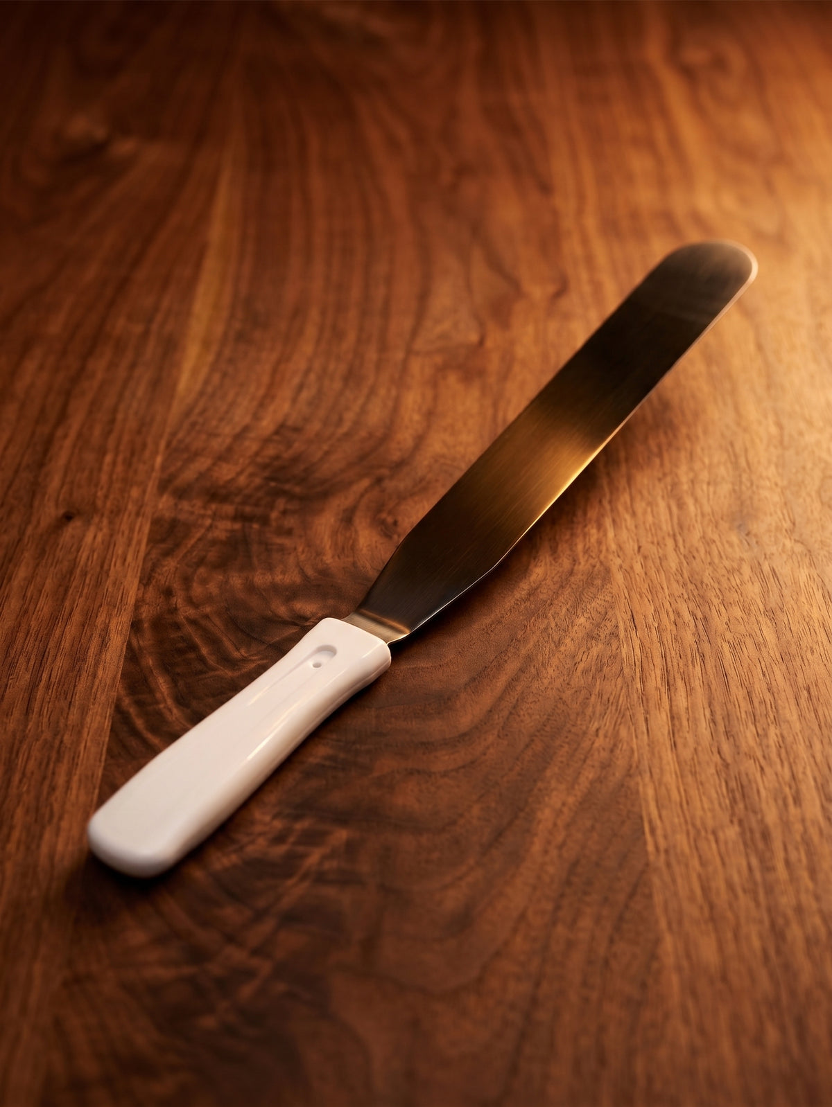 A 12" metal crepe spatula with a white plastic handle resting diagonally on a warm, dark wood grain surface.