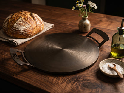 A round dark nitrogen infused carbon steel 14" 726 Pan with side handles sits on a rustic wooden table, styled with a loaf of bread, a small vase of flowers, a bottle of oil, and a bowl of salt.